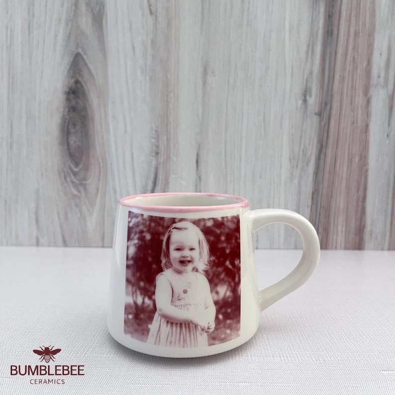Puede incluir: Una taza de cer&aacute;mica blanca con un borde rosa y una impresi&oacute;n fotogr&aacute;fica monocrom&aacute;tica de color marr&oacute;n rojizo de un ni&ntilde;o sonriente en el frente. La taza tiene un asa curva y un acabado liso, mostrando una imagen clara de una persona.