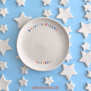 May include: White ceramic plate with the words "happy birthday" in rainbow colors and the name "Stetson" in rainbow colors. The plate is surrounded by white ceramic stars.