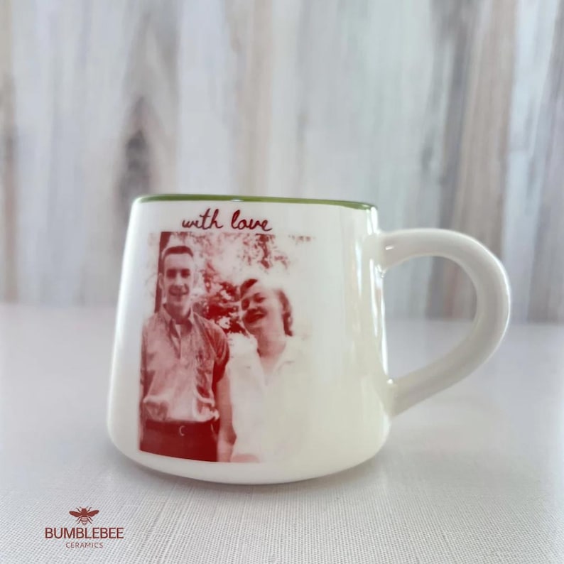 Puede incluir: Una taza de cer&aacute;mica blanca con borde verde que presenta una impresi&oacute;n fotogr&aacute;fica en tonos rojos de dos personas y el texto manuscrito "with love" sobre la imagen. La taza tiene un asa curva y un acabado exterior liso y brillante.