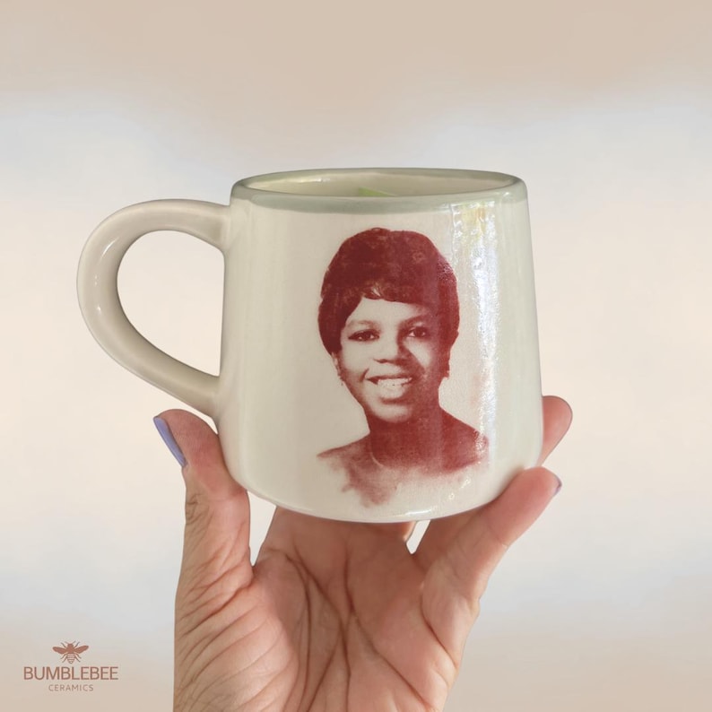 Puede incluir: Una taza de cer&aacute;mica de color crema con un retrato de color marr&oacute;n rojizo de una persona sonriente con el pelo corto impreso en el frente. La taza tiene un acabado liso y brillante, un asa redondeada a la izquierda y una forma cil&iacute;ndrica.