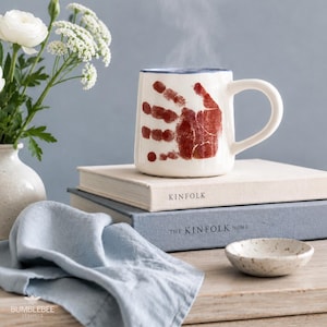 Puede incluir: Una taza de cer&aacute;mica blanca con un dise&ntilde;o de huella de mano de color marr&oacute;n rojizo en el frente, apoyada sobre dos libros apilados titulados Kinfolk y The Kinfolk Home. La taza tiene un borde azul y un asa curva, con vapor saliendo de ella.