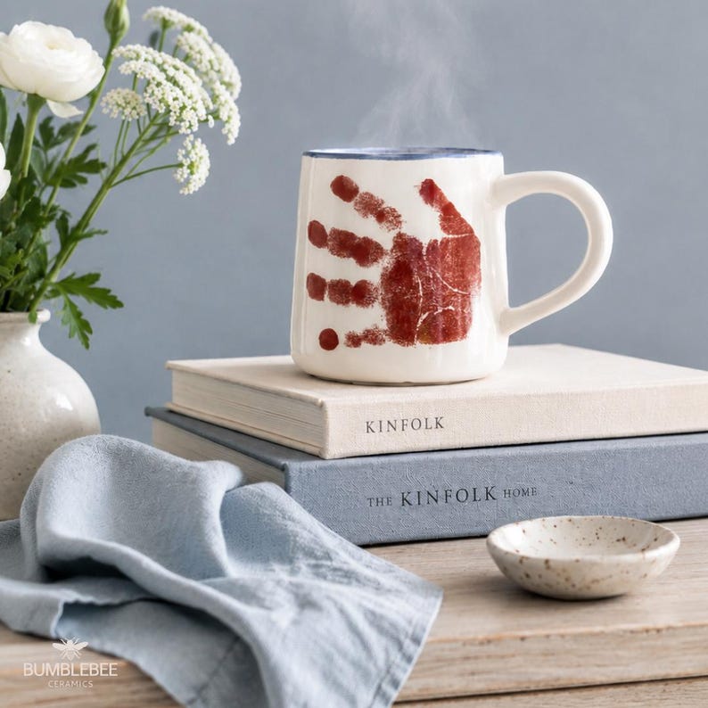 Puede incluir: Una taza de cer&aacute;mica blanca con un dise&ntilde;o de huella de mano de color marr&oacute;n rojizo en el frente, apoyada sobre dos libros apilados titulados Kinfolk y The Kinfolk Home. La taza tiene un borde azul y un asa curva, con vapor saliendo de ella.