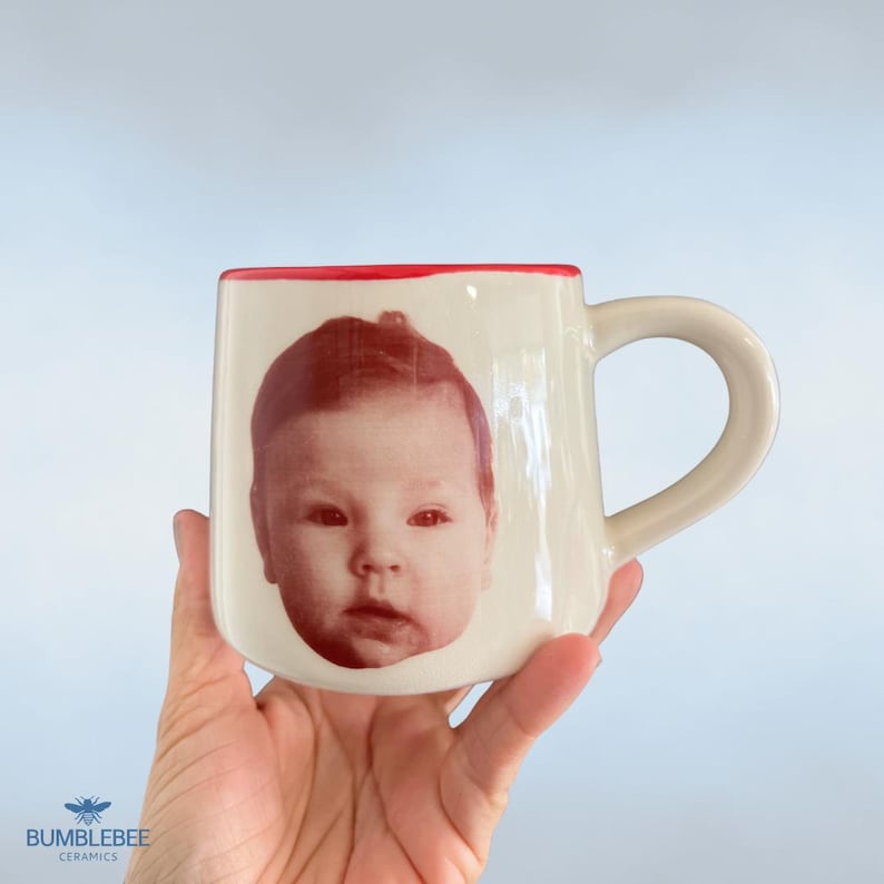 Puede incluir: Una taza de cer&aacute;mica de color crema con una impresi&oacute;n fotogr&aacute;fica en tonos rojos de la cara de un beb&eacute; en la parte frontal. La taza tiene un asa redondeada y un borde rojo distintivo en la parte superior. El acabado es brillante.