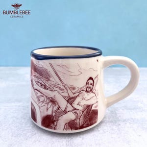 Puede incluir: Una taza de cer&aacute;mica color crema con borde azul oscuro, que presenta un dibujo lineal de color granate de una persona sentada en un barco. La ilustraci&oacute;n muestra a una persona con barba, vistiendo pantalones cortos, relaj&aacute;ndose en la cubierta.