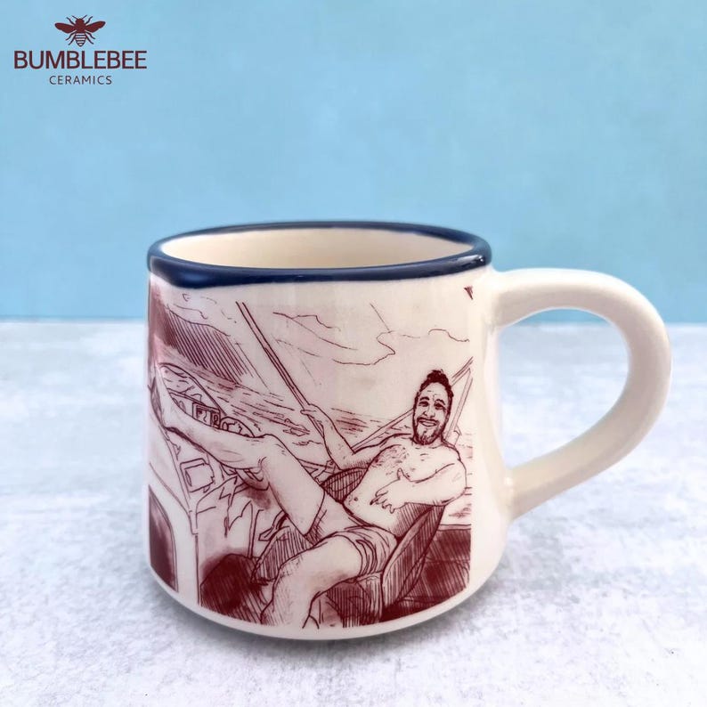 Puede incluir: Una taza de cer&aacute;mica color crema con borde azul oscuro, que presenta un dibujo lineal de color granate de una persona sentada en un barco. La ilustraci&oacute;n muestra a una persona con barba, vistiendo pantalones cortos, relaj&aacute;ndose en la cubierta.