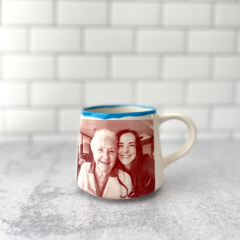 Puede incluir: Una taza de cer&aacute;mica con cuerpo redondeado y asa, que presenta una impresi&oacute;n fotogr&aacute;fica en tono sepia de dos personas sonrientes en el frente. El borde de la taza est&aacute; pintado con una banda azul brillante, contrastando con el color blanco roto.
