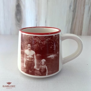 Puede incluir: Una taza de cer&aacute;mica blanca con borde rojo y asa, que presenta una impresi&oacute;n fotogr&aacute;fica en tono sepia de dos personas en el agua. La taza tiene una base redondeada y un acabado liso, con el texto BUMBLEBEE CERAMICS impreso en color rojo.