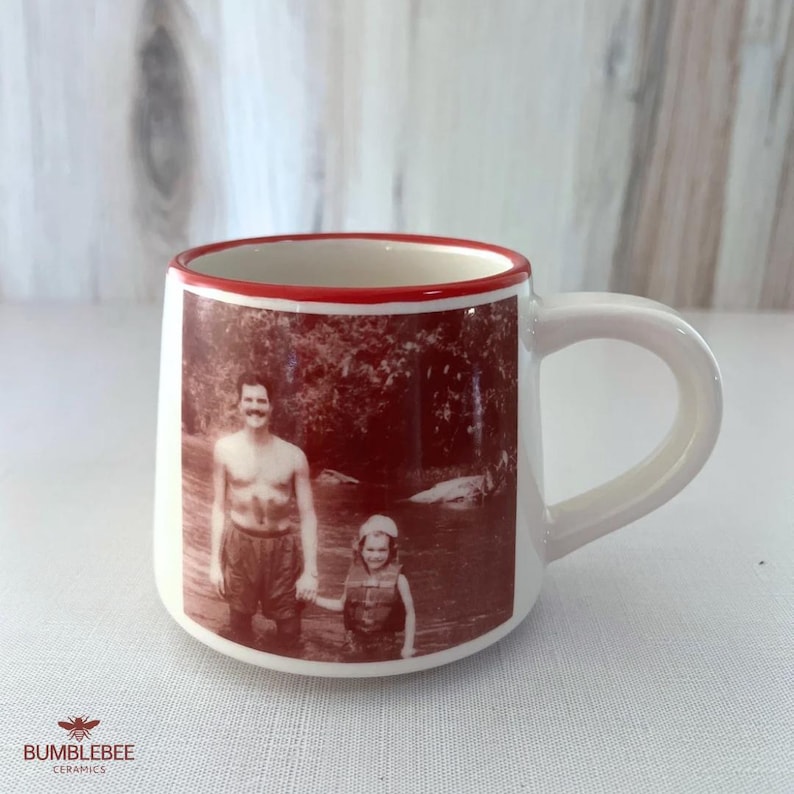 Puede incluir: Una taza de cer&aacute;mica blanca con borde rojo y asa, que presenta una impresi&oacute;n fotogr&aacute;fica en tono sepia de dos personas en el agua. La taza tiene una base redondeada y un acabado liso, con el texto BUMBLEBEE CERAMICS impreso en color rojo.