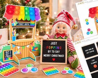 Puede incluir: Un puesto de fidget de cartón con techo arcoíris, un letrero que dice FIDGET BOOTH y una pizarra que dice BUY 2 GET 1 FREE. Una muñeca con gorro rojo y blanco sostiene un tablero de letras negro que dice JUST POPPING BY TO SAY MERRY CHRISTMAS.