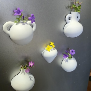 Puede incluir: Varios jarrones magnéticos blancos, impresos en 3D, de diversas formas, cada uno con flores coloridas. Los jarrones están adheridos a una superficie gris, creando un arreglo floral decorativo. Las flores incluyen flores moradas, rosas y amarillas.