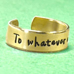 May include: A gold-coloured adjustable ring with the phrase "To whatever!" in black lettering. The ring is set against a green background. The ring is a piece of jewellery.