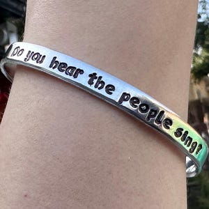 May include: A silver cuff bracelet with the engraved text "Do you hear the people sing?". The bracelet has a curved design and is worn on a wrist. The text is in a bold, sans-serif font.