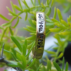 May include: A gold-colored ring with the words "as you wish" engraved in black lettering. The ring is set against a backdrop of green foliage and red berries, creating a natural and organic aesthetic.