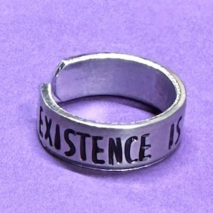 Puede incluir: Un anillo plateado con las palabras "EXISTENCE IS" grabadas alrededor de la banda. El anillo está sobre un fondo morado.