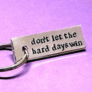 以下が含まれることがあります： 「don't let the hard days win」というフレーズが黒い文字で刻印された長方形のシルバーキーホルダー。キーホルダーはシルバーの二重リングに取り付けられています。背景は紫色の無地です。