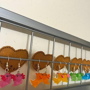 May include: A display of colorful axolotl earrings hanging from a metal rack. The earrings are in various colors, including pink, orange, yellow, green, blue, and purple. Each axolotl is suspended from a silver hook, and displayed on a brown heart-shaped card.