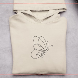 May include: A cream-colored hoodie with a black line drawing of a butterfly on the front. The butterfly design is minimalist, with a single continuous line forming the wings and body. The hoodie has a hood and a kangaroo pocket.