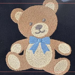 May include: Embroidered teddy bear patch with a light brown body, tan accents, and a blue bow tie. The bear has a stitched design with a smiling face and is set against a dark blue background.