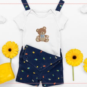 May include: A white baby onesie with a brown teddy bear design and a blue bow, paired with dark blue denim shorts featuring colorful patterns. Yellow rubber boots and sunflowers complete the playful ensemble.