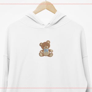 May include: White hoodie with a brown teddy bear embroidered on the front. The teddy bear has a blue bow tie. The hoodie is hanging on a wooden hanger against a white background.