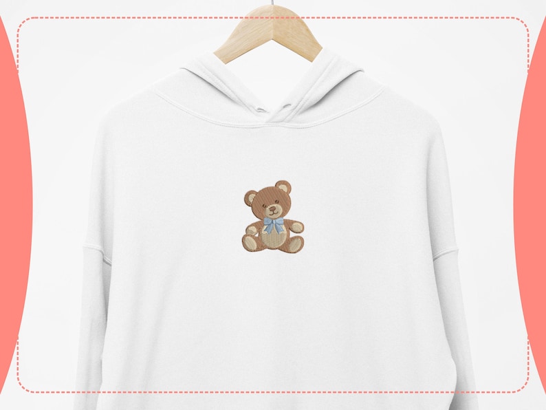 May include: White hoodie with a brown teddy bear embroidered on the front. The teddy bear has a blue bow tie. The hoodie is hanging on a wooden hanger against a white background.