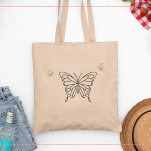 May include: Beige tote bag featuring a large black butterfly outline in the center, with two smaller butterfly outlines on either side. The bag has two handles and is made of a light-colored fabric. The design is simple and elegant.