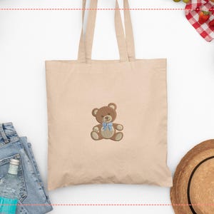 May include: Beige tote bag with a brown teddy bear embroidery wearing a blue bow tie. The bag has two handles and is made of a light-colored fabric. The image also shows a pair of blue jeans, a straw hat, and a small bowl of strawberries.