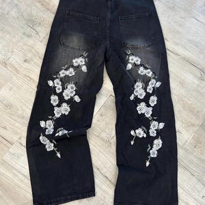 Op de afbeelding: Zwarte spijkerbroek met wijde pijpen en een hoge taille. De achterkant van de broek heeft twee zakken en gedetailleerd wit bloemenborduursel dat zich uitstrekt van de bovenbenen tot aan de onderkant van de broekspijpen.