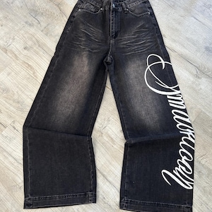 Peut inclure: Jean large à taille haute dans un délavage noir usé avec des détails de moustaches au niveau des hanches. La jambe gauche présente un grand texte en écriture cursive blanche courant verticalement sur le côté. Fermeture à bouton et zip.