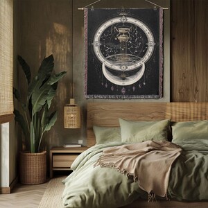 May include: A black tapestry with a celestial design, featuring a central pillar, constellations, and a crescent moon. The tapestry is hanging above a bed with green bedding and a tan throw. A large potted plant and a woven lamp are also visible.
