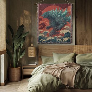 May include: A woven wall tapestry featuring a Godzilla-like creature against a red sun and ocean waves. The artwork is in shades of teal, blue, and red, with a fringed border. The tapestry is hanging above a bed with green bedding.