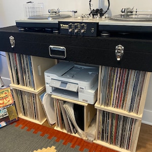 May include: A wooden record storage unit with six shelves, each filled with vinyl records. A silver printer sits on top of the unit, and two turntables and a mixer are on top of the unit. The unit is black with a silver handle.