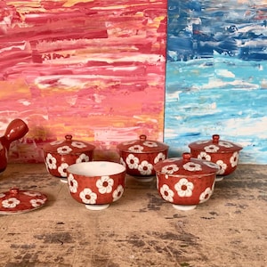 May include: A red ceramic tea set with white floral designs. The set includes a teapot, several covered bowls, and a teacup. The background features colorful abstract paintings. The tea set is likely for serving tea.