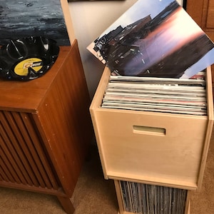 May include: A wooden record storage unit with a light wood finish. The unit has two sections, one for storing records and the other for displaying a record cover. The unit is sitting on a wooden floor.