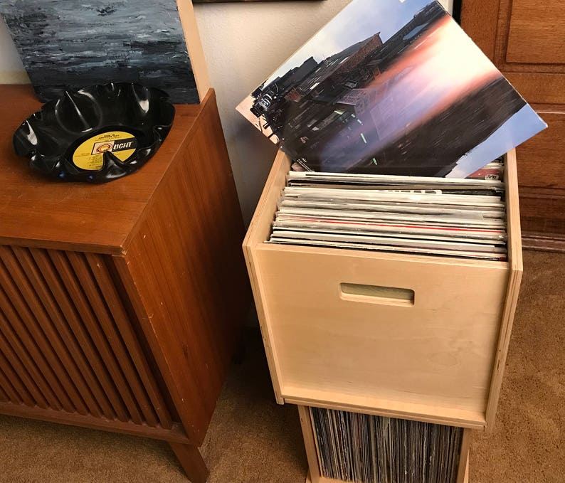 May include: A wooden record storage unit with a light wood finish. The unit has two sections, one on top of the other. The top section has a door with a handle. The bottom section is open and has a stack of vinyl records inside. The unit is sitting on a carpeted floor.