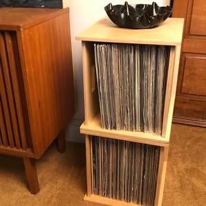 May include: A two-tiered wooden record storage unit with a black vinyl record player on top. The unit is filled with vinyl records.