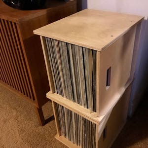 May include: Two stacked wooden record storage crates with a light wood finish. The crates are filled with vinyl records.