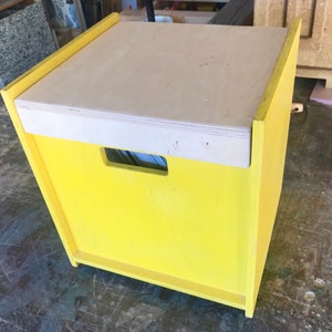 May include: A yellow wooden storage box with a light wood lid and a cut-out handle. The box is sitting on a gray surface.