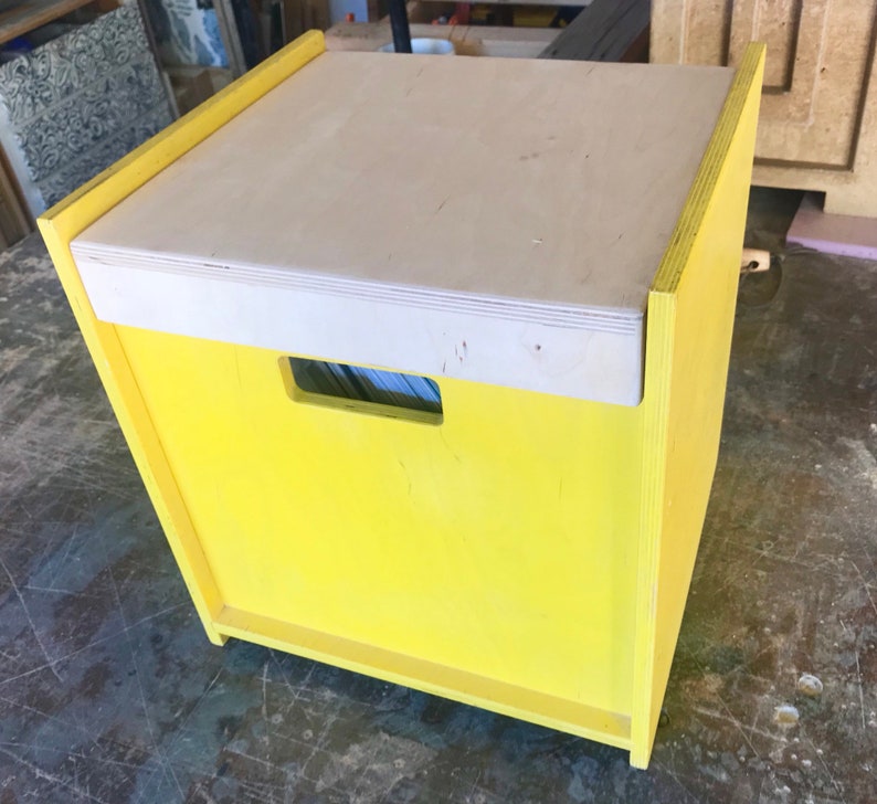 May include: A yellow wooden storage box with a light wood lid and a cut-out handle. The box is sitting on a gray surface.