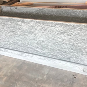 Op de afbeelding: Een grote, rechthoekige, grijze betonnen plantenbak met een ruw, getextureerd oppervlak. De plantenbak heeft een licht verhoogde rand rond de bovenrand.