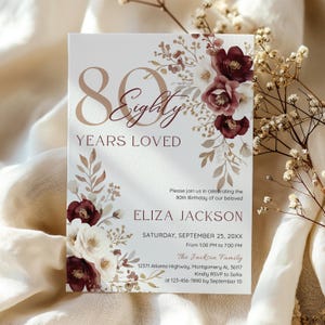 May include: A white invitation card with the text "80 Years Loved" in a copper color. The card is decorated with burgundy and white floral arrangements. The invitation is for Eliza Jackson's 80th birthday, with event details and RSVP information.
