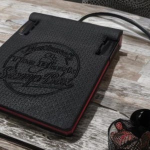 May include: Black rectangular stomp box with a textured surface and red trim. The box has the text "The Stomp Box" in a circular design. A silver-colored connector is attached to the box with a black cable. A guitar pick is also visible.