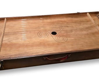 May include: A wooden hammered dulcimer with numerous strings stretched across a light brown soundboard. The instrument is housed in a dark brown rectangular case with a handle and metal latches. A Celtic knot design is etched into the wood.