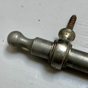 May include: A silver-toned metal cabinet pull featuring a cylindrical bar, a rounded ball-shaped end, and a decorative mounting post with an attached threaded screw. The hardware exhibits a metallic finish with visible signs of wear and oxidation.