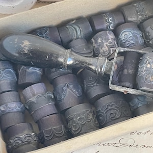 Pode incluir: Uma coleção de selos antigos, roxos escuros, com desenhos ornamentados, guardados numa caixa de cartão. Os selos têm formato cilíndrico e são feitos de um material escuro. Uma pega de selo com uma pega de madeira preta e detalhes em metal prateado repousa no topo.