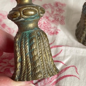 May include: A brass tassel-shaped object with a rope-textured base and a rounded, decorative top. The metal surface displays a weathered patina with visible green oxidation in the recessed areas, showing a detailed, molded corded design.