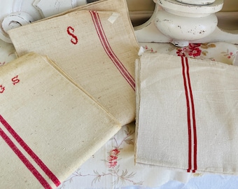 May include: Three vintage linen tea towels with red stripes and embroidered letters. The towels are folded and displayed on a floral patterned surface.