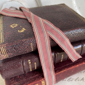 May include: A pile of antique books with dark burgundy leather covers. A striped ribbon in red, grey, and cream is tied around the books. Gold lettering is visible on the spines and covers, giving a vintage feel.