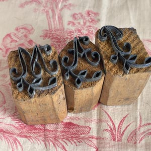 Könnte beinhalten: Drei alte Letterpress-Druckblöcke mit dem Buchstaben "A" in einer dekorativen Schrift. Die Metallbuchstaben sind auf rechteckigen Holzblöcken montiert. Die Blöcke sind auf einem rosa-weiß geblümten Stoff angeordnet.