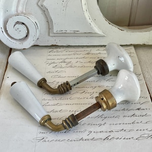 Può includere: Due maniglie per porte vintage in porcellana bianca con dettagli in ottone. Le maniglie hanno un design classico e sono poste su uno sfondo di scrittura antica. L'hardware è probabilmente per un armadio o una porta.
